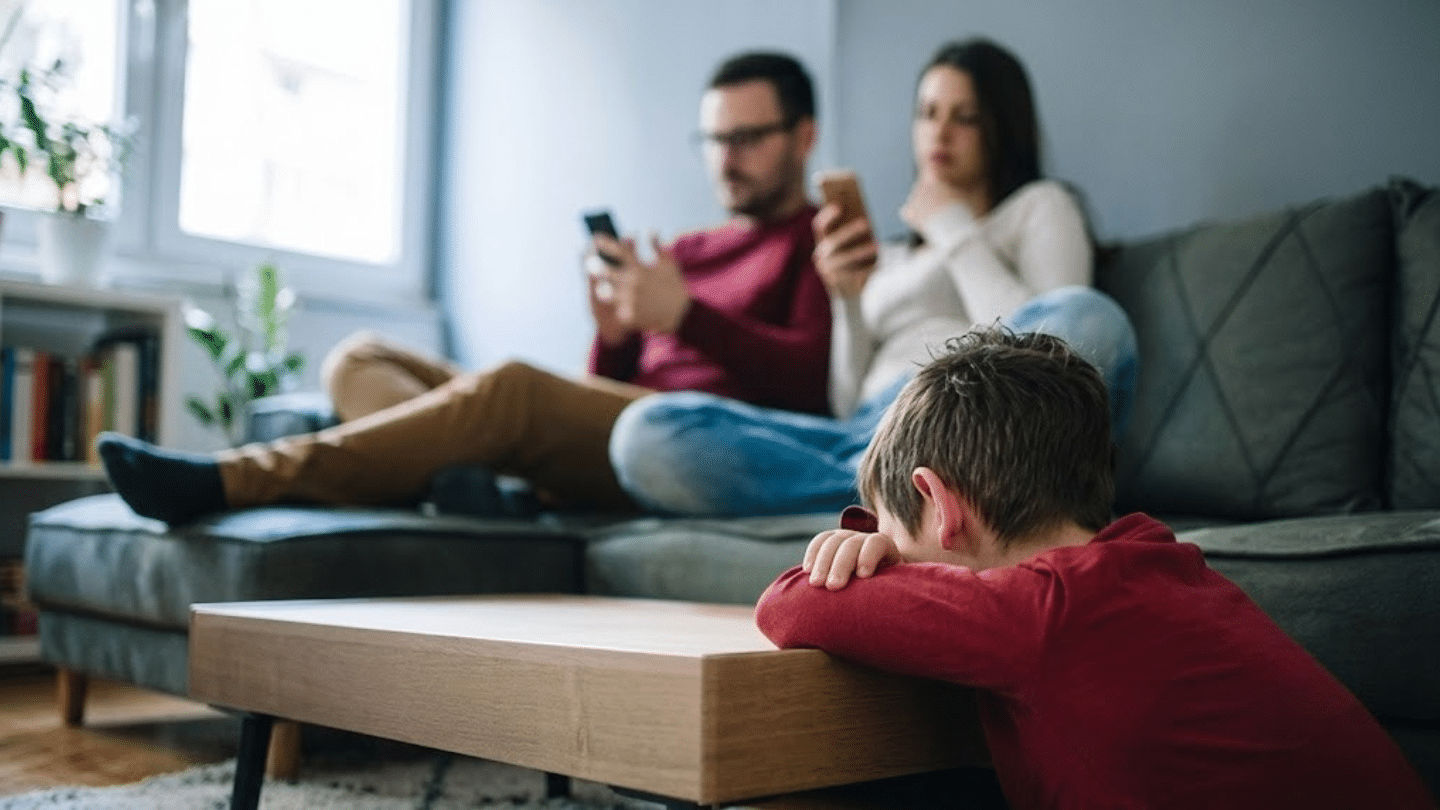 a young boy leans sadly on a table while his parents sit blurred in the background looking at their phones