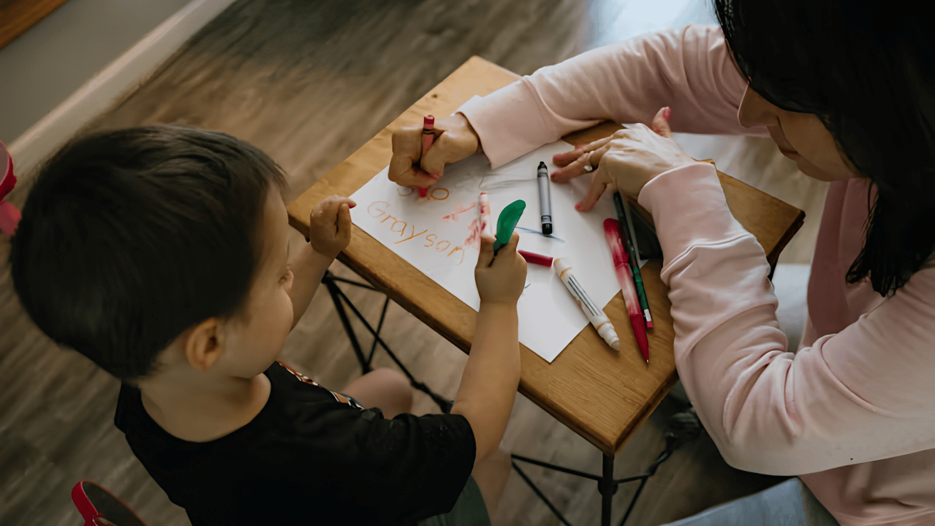 adult helping a young child practice writing with colorful markers at a table