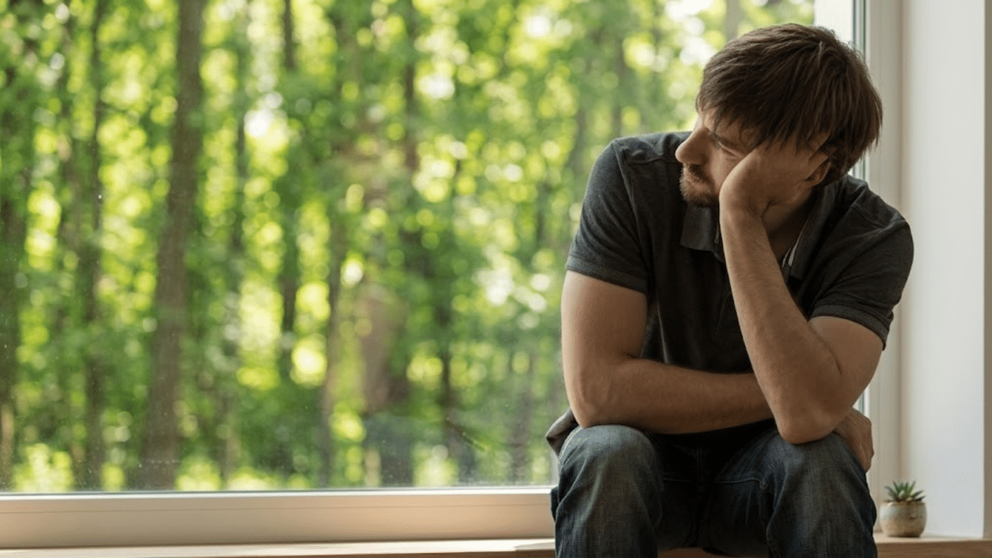 pensive man sitting by a window resting his face in his hand while looking out at a bright green forest