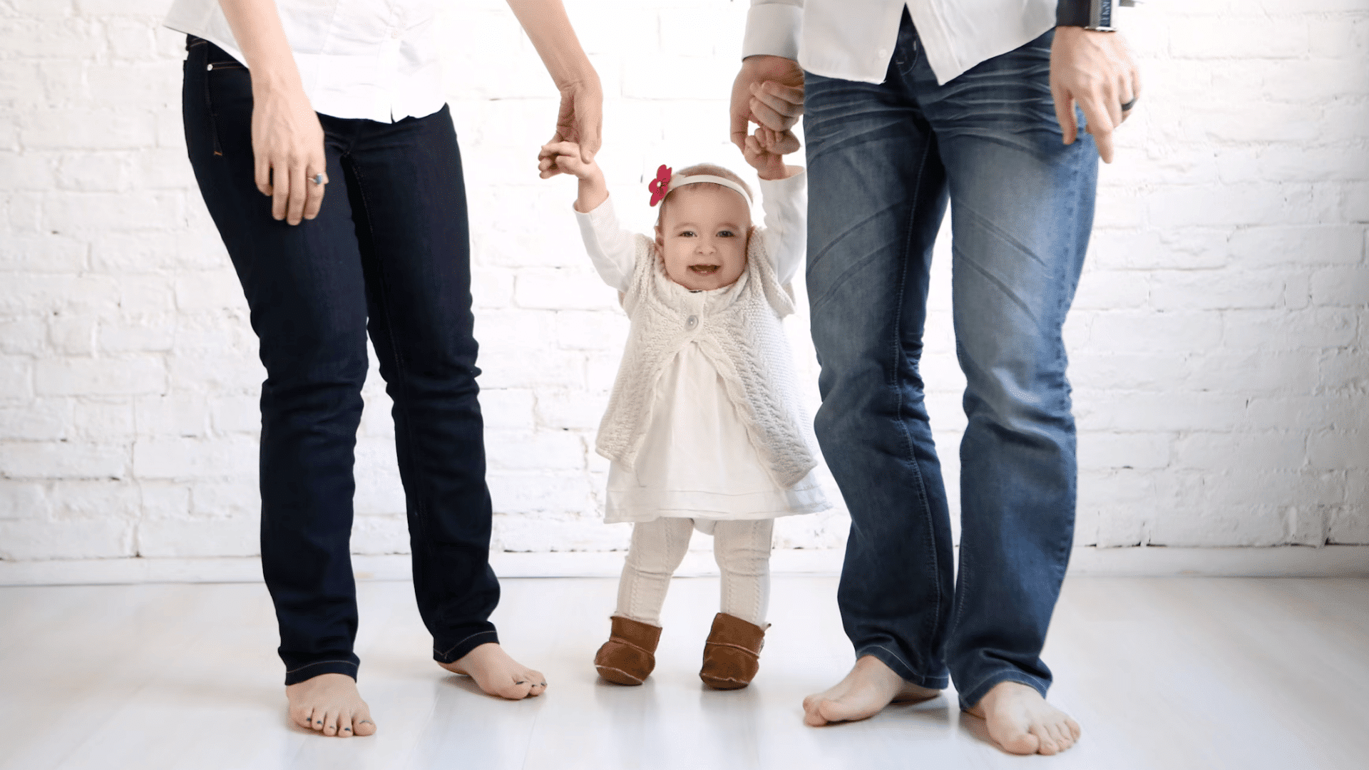 smiling toddler takes early steps while being supported by two adults holding both hands indoors