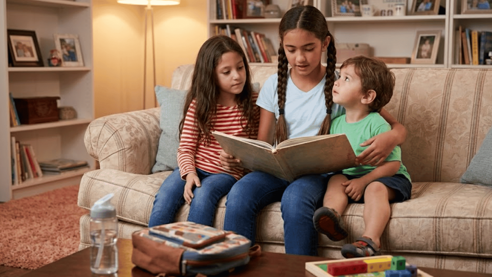 three children sitting together on a couch reading a book in a cozy living room with bookshelves and family photos behind them