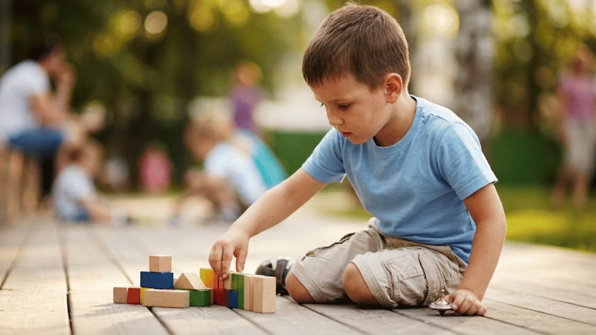 young boy kneeling on a wooden deck stacking colorful building blocks while children play blurred in a park
