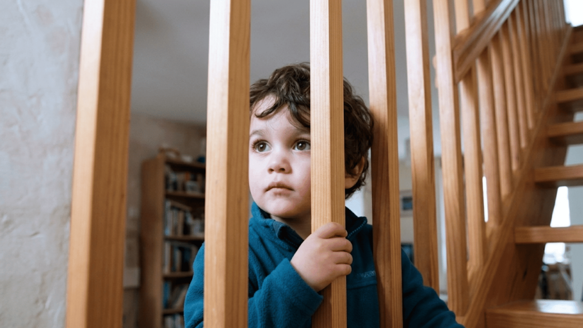 young child holding wooden stair railings while standing indoors near a staircase with a quiet thoughtful look