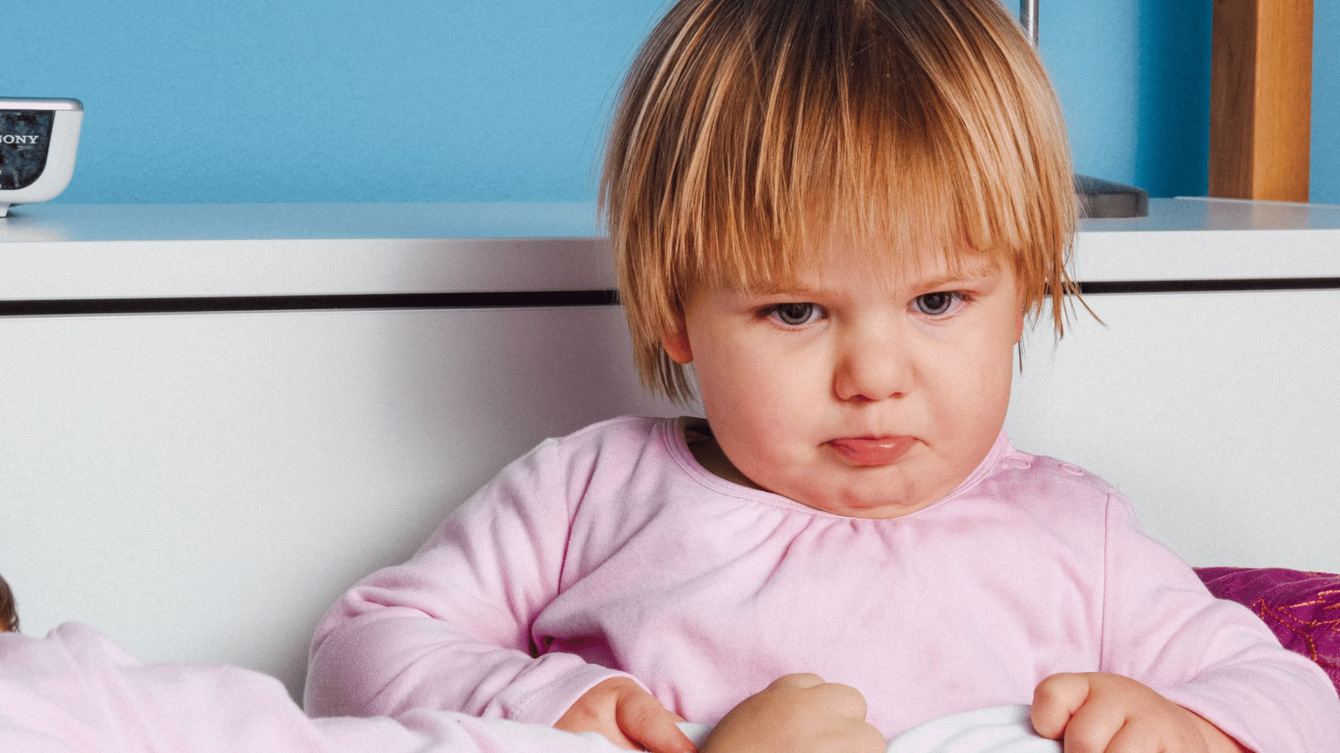 young child in pink pajamas sits on a bed with a sad expression looking down while holding a blanket