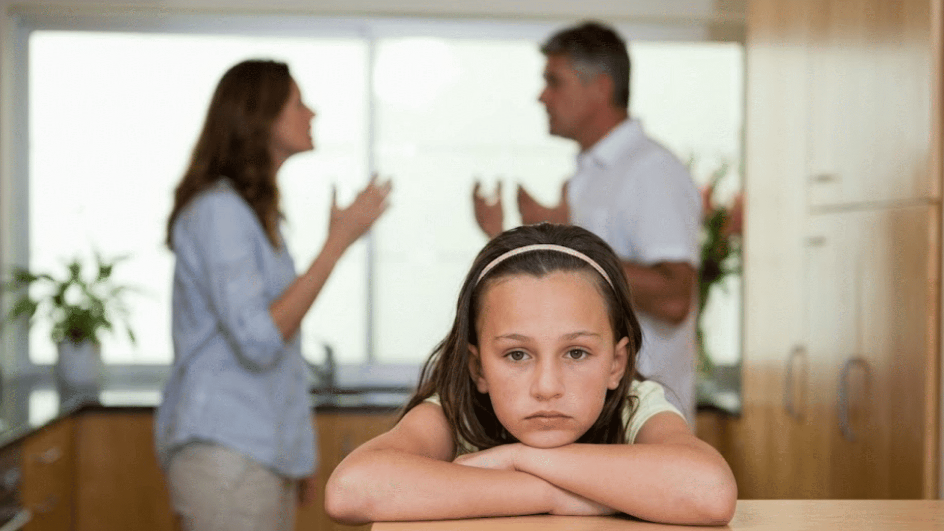 young girl resting her arms on a table looking sad while parents argue in the background of a bright kitchen scene