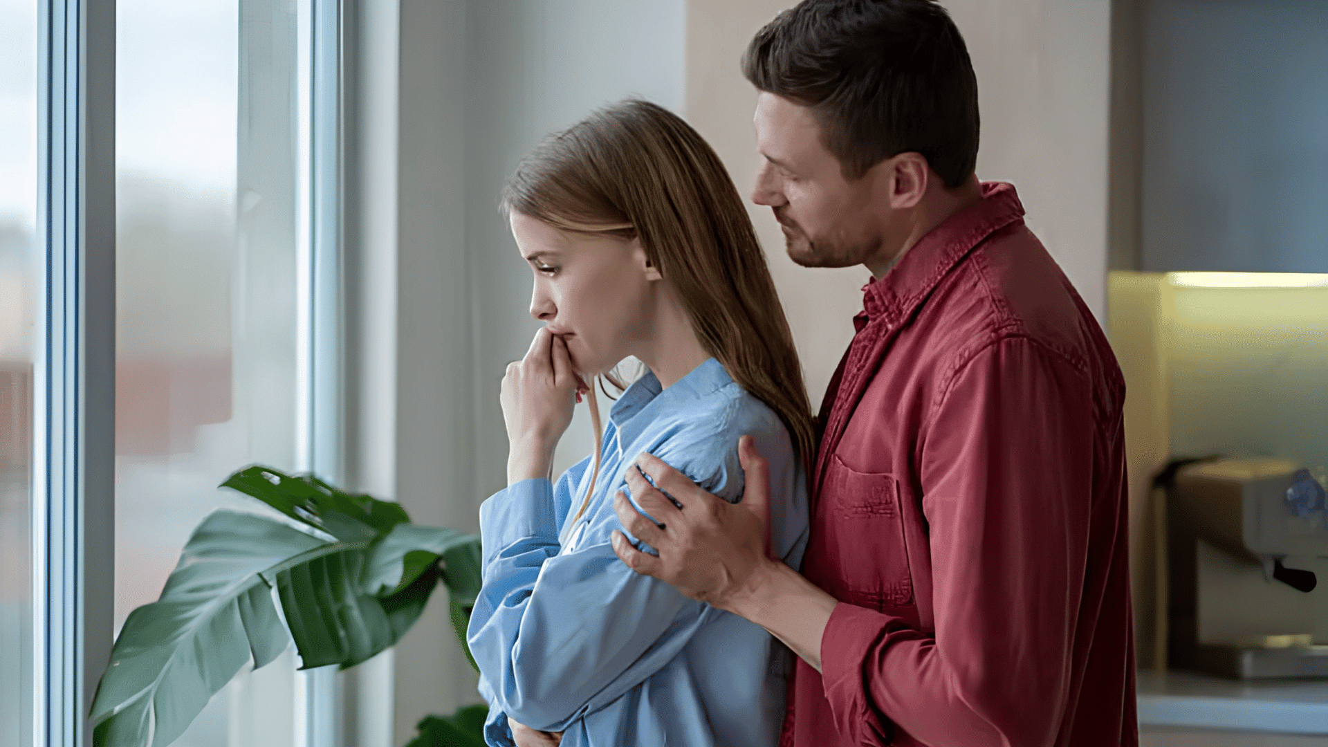 a man comforts a worried woman as they stand by a window looking outside