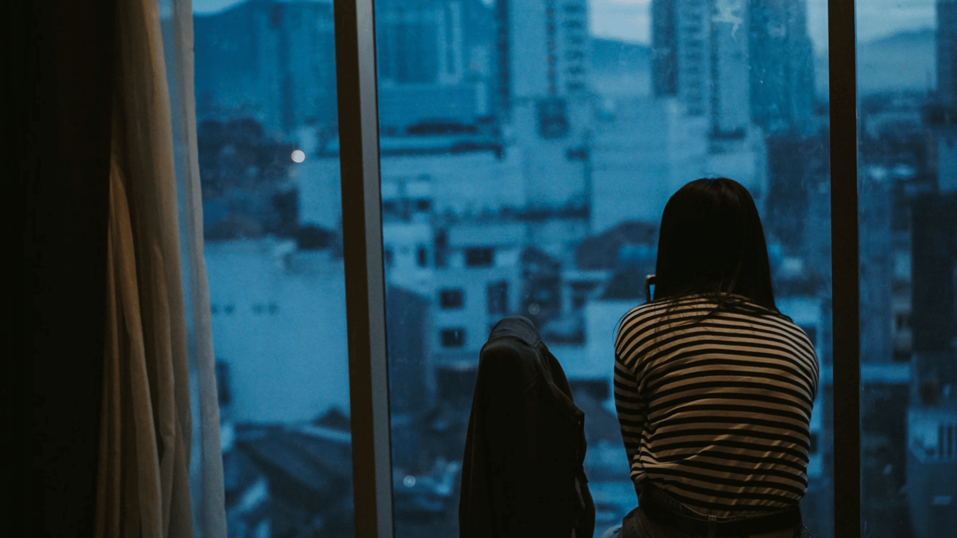 a person sits alone by a large window at dusk looking out over a blue city skyline while holding a phone