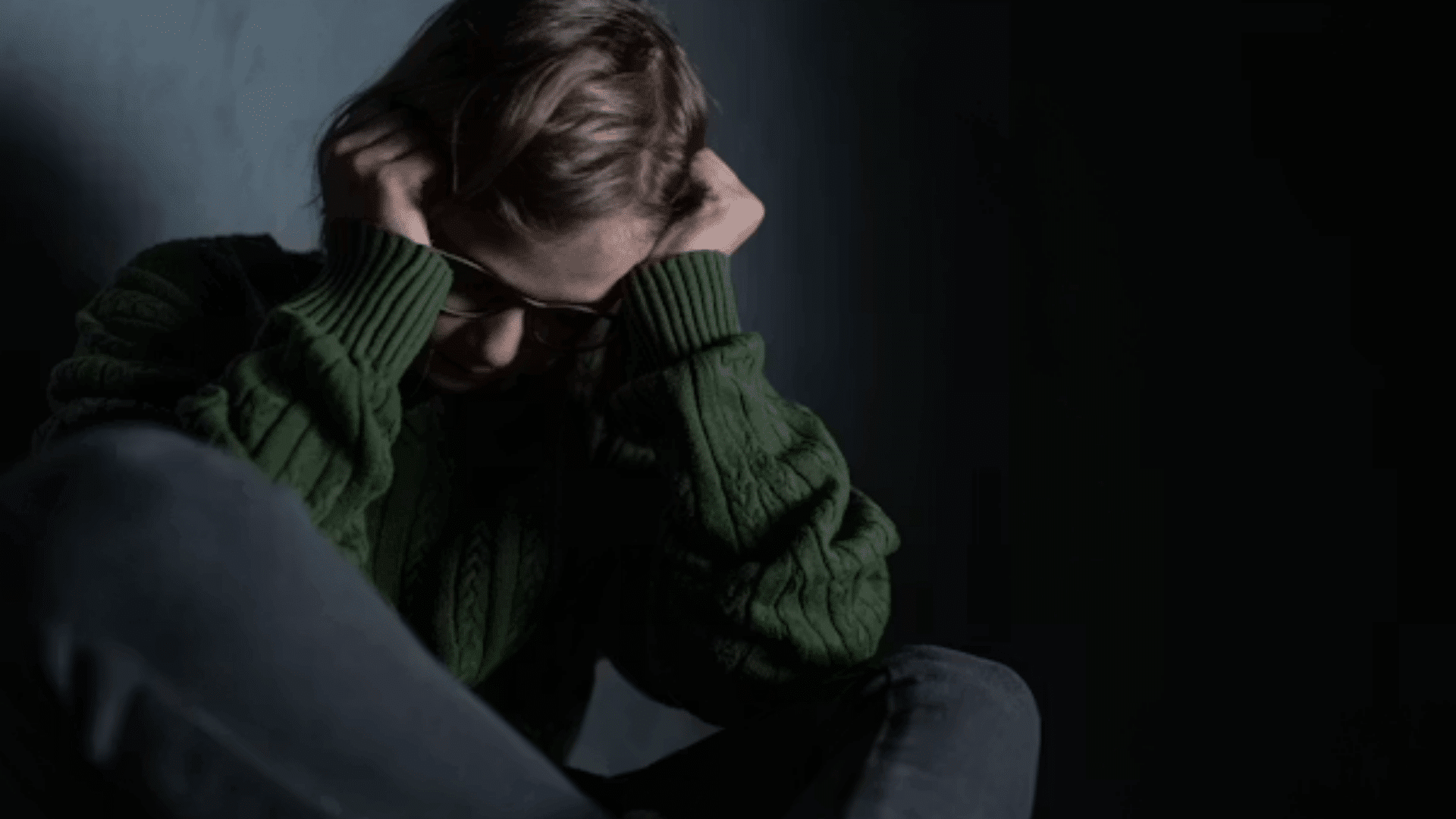 a person sits on the floor with hands over ears appearing distressed in a dark room