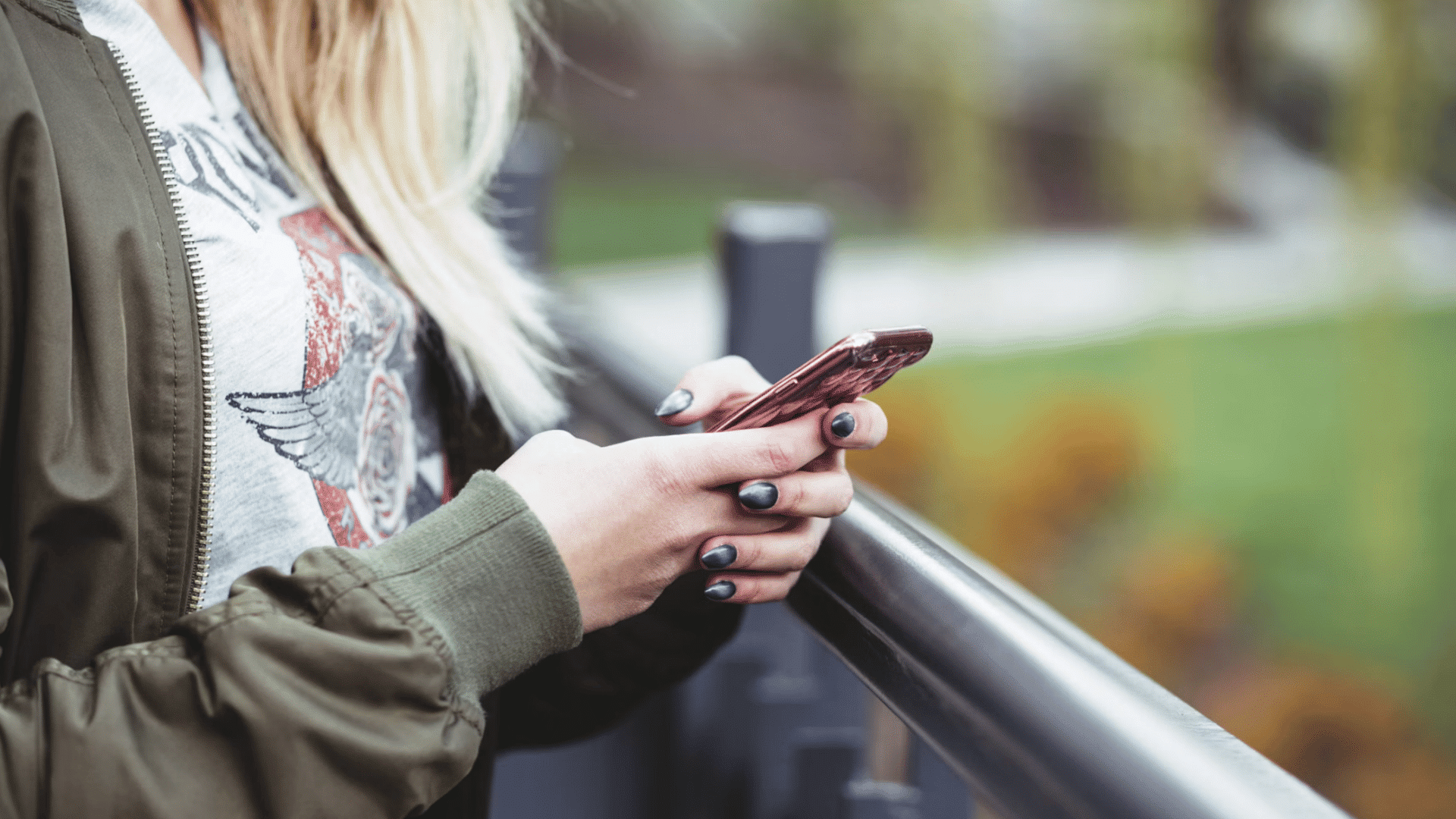 a person standing by a railing uses a pink phone with a softly blurred green outdoor background
