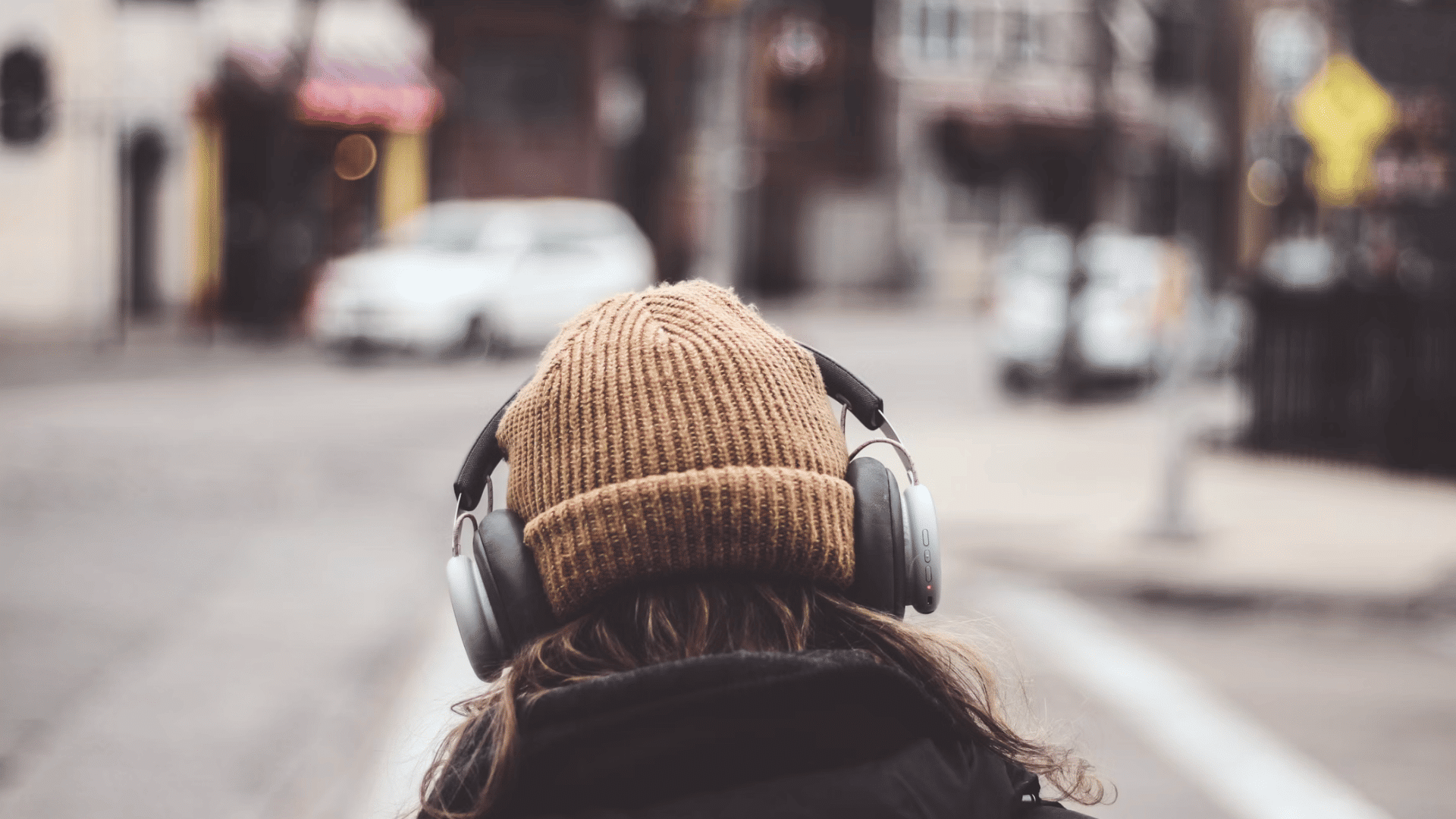 a person wearing headphones and a knit hat walks through a blurred city street from behind