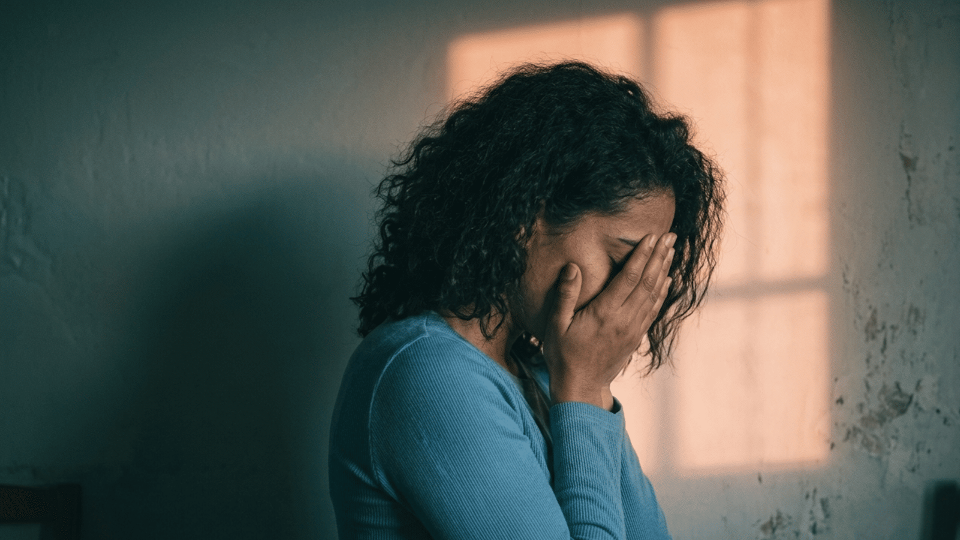 a sad woman covering her face while sitting alone in a dim room with soft window light