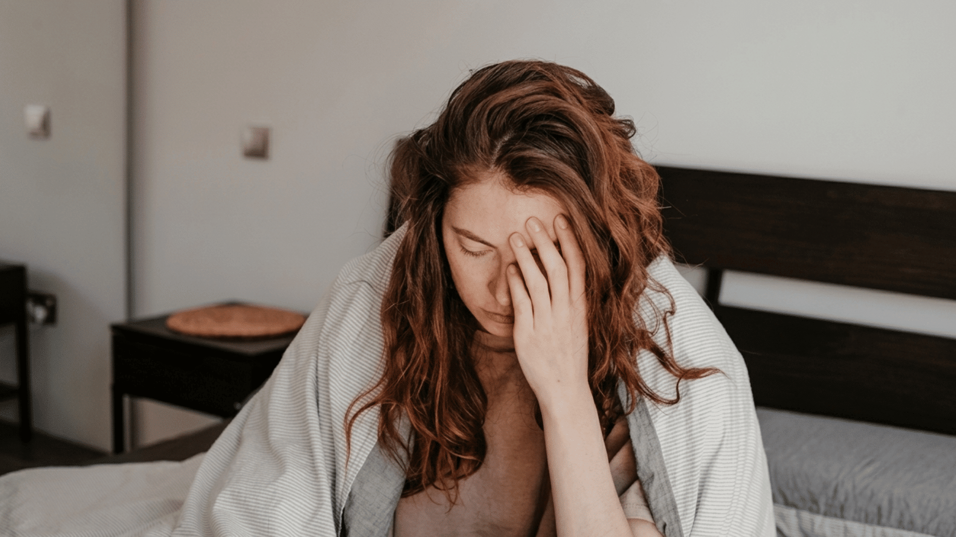 a tired woman wrapped in a blanket sitting on bed holding her forehead in discomfort