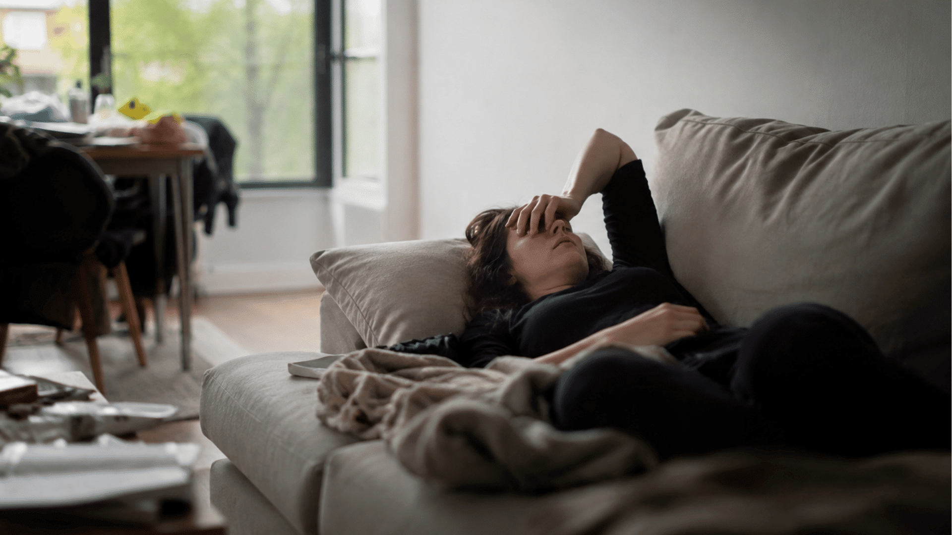a woman lying on a sofa with her hand over her face in a living room beside a blanket and scattered items