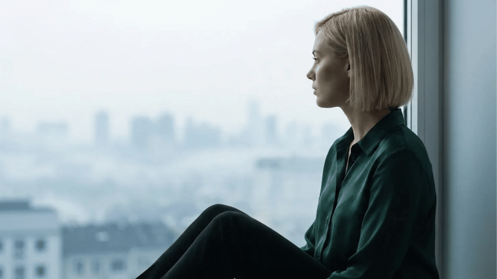 a woman sitting by a large window gazing toward a distant city skyline with a thoughtful expression indoors