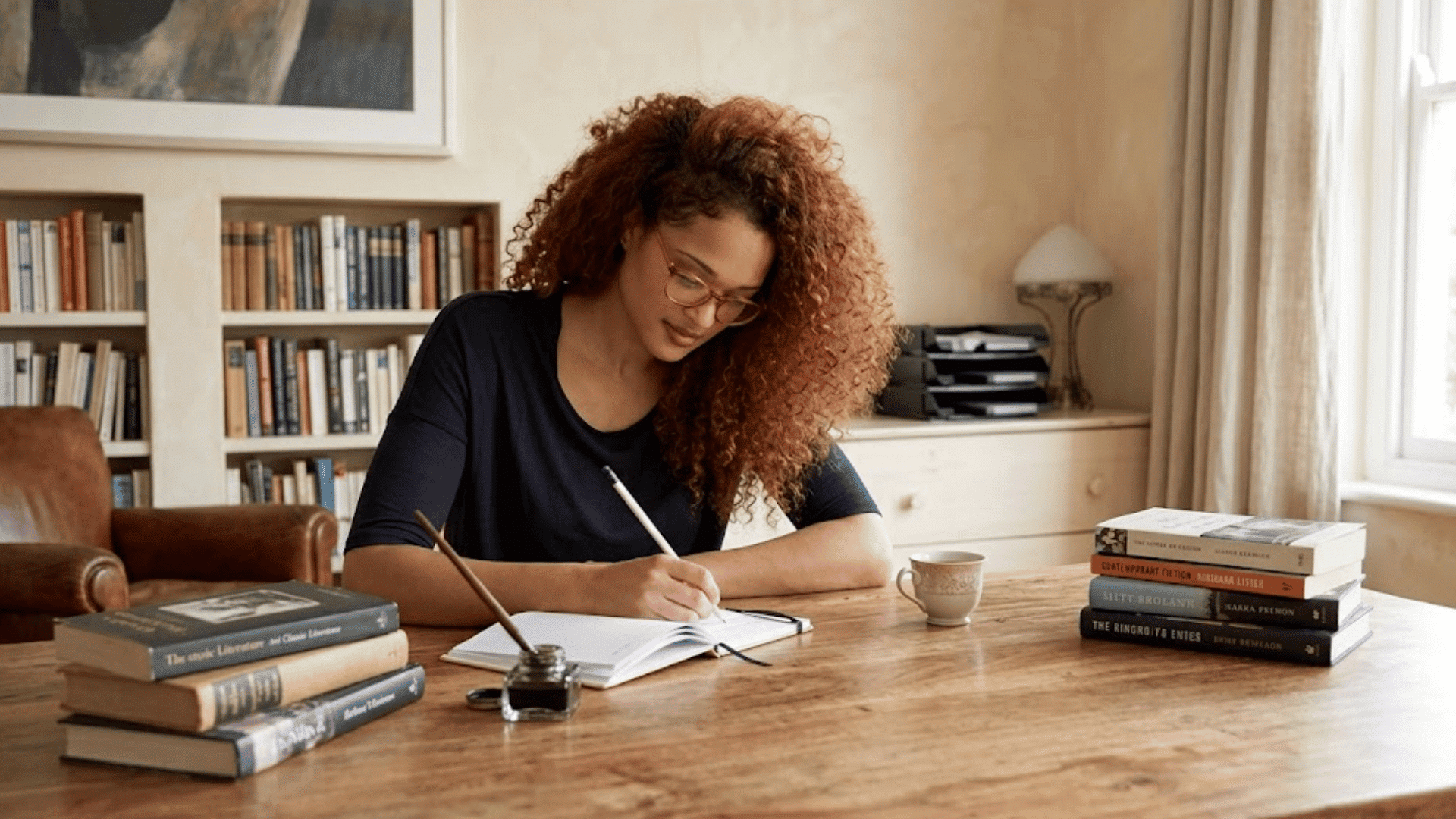 a woman with curly hair writes in a notebook at a wooden table surrounded by books in a cozy home study room