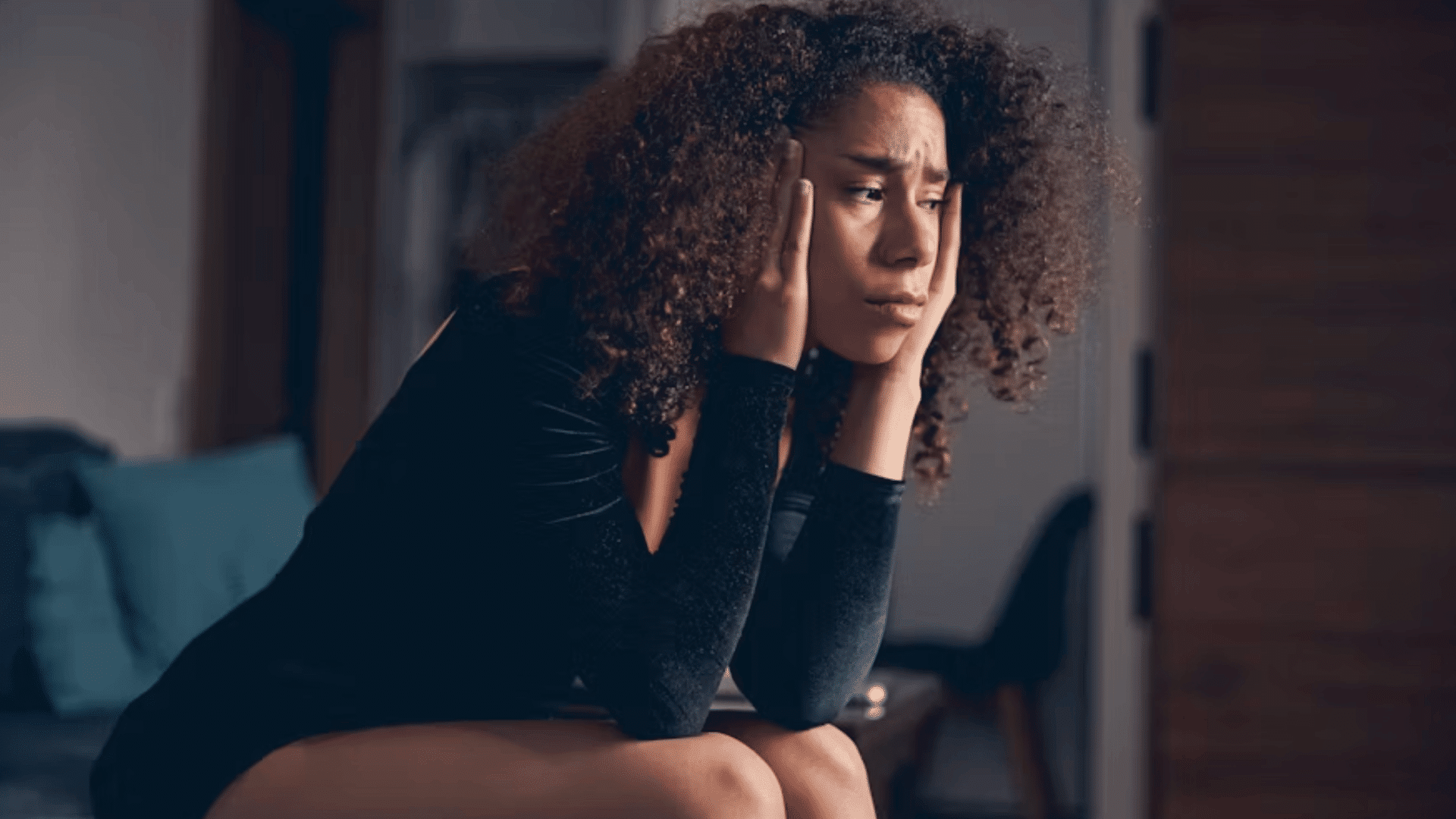 a worried woman sits on a couch holding her head in her hands while looking away in a dim room