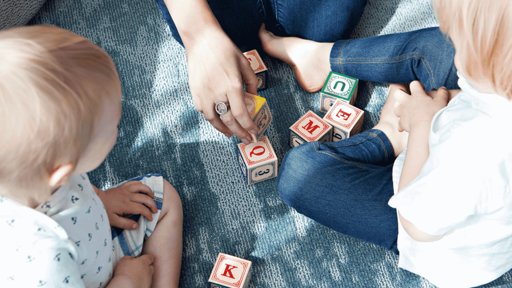 adult and two young children sitting on a carpet stacking colorful alphabet blocks together during indoor playtime (1)