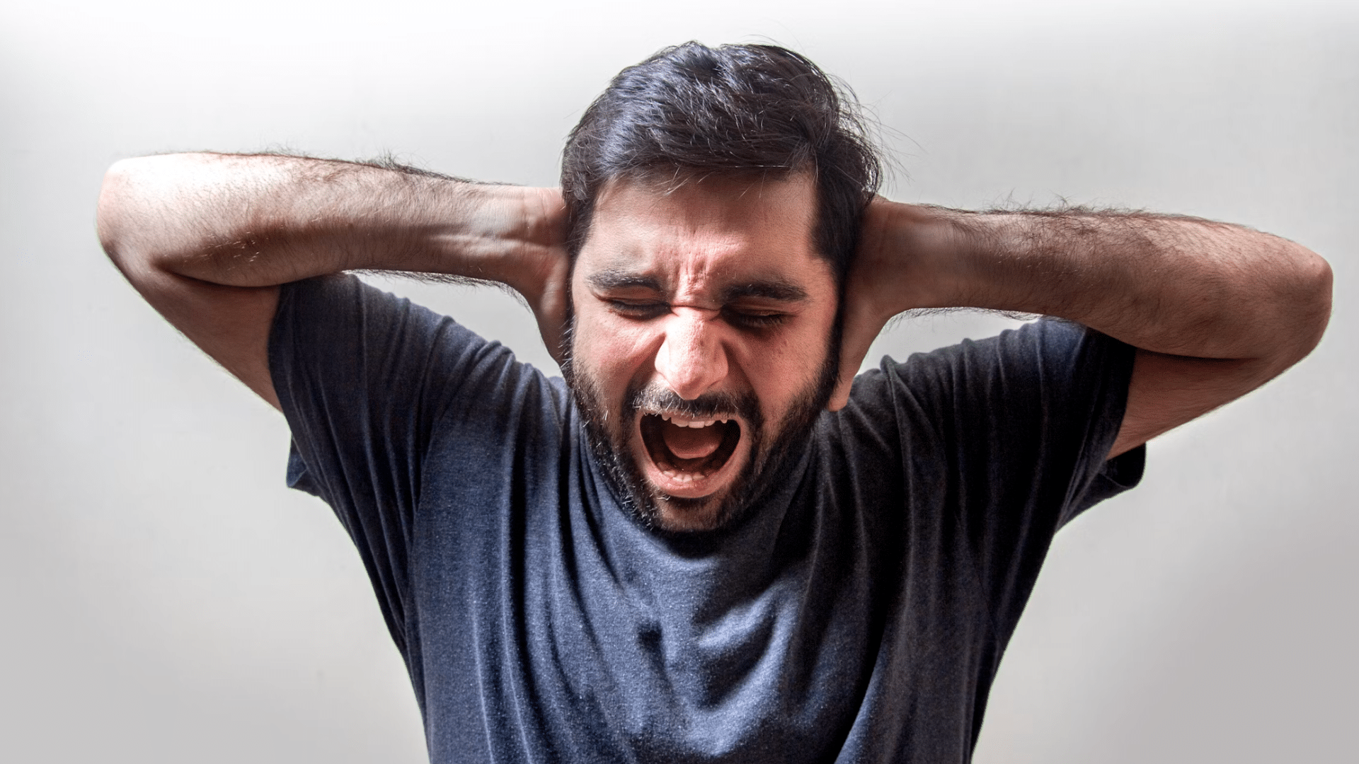 bearded man shouts with eyes closed and hands covering his ears against a plain light background
