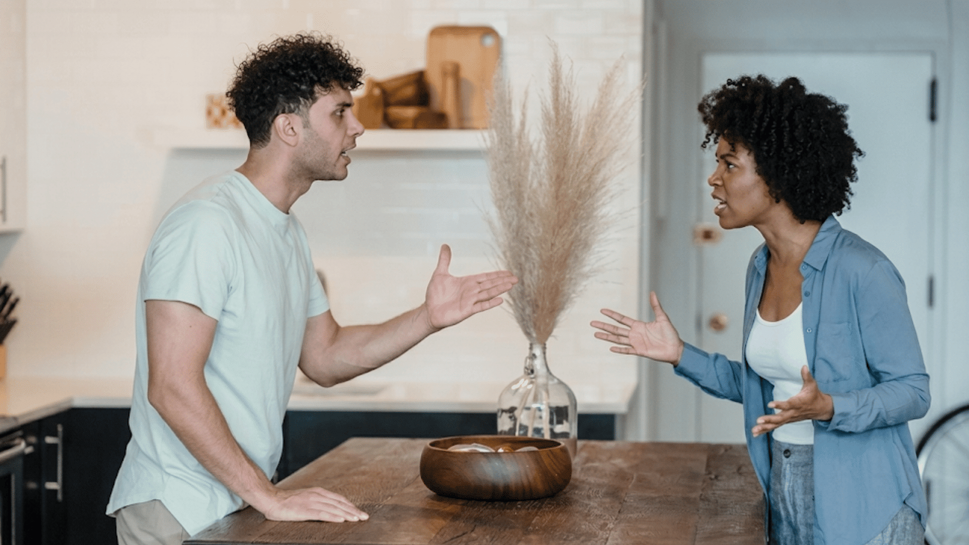 couple argues across a kitchen island with tense gestures during a disagreement at home near a wooden bowl