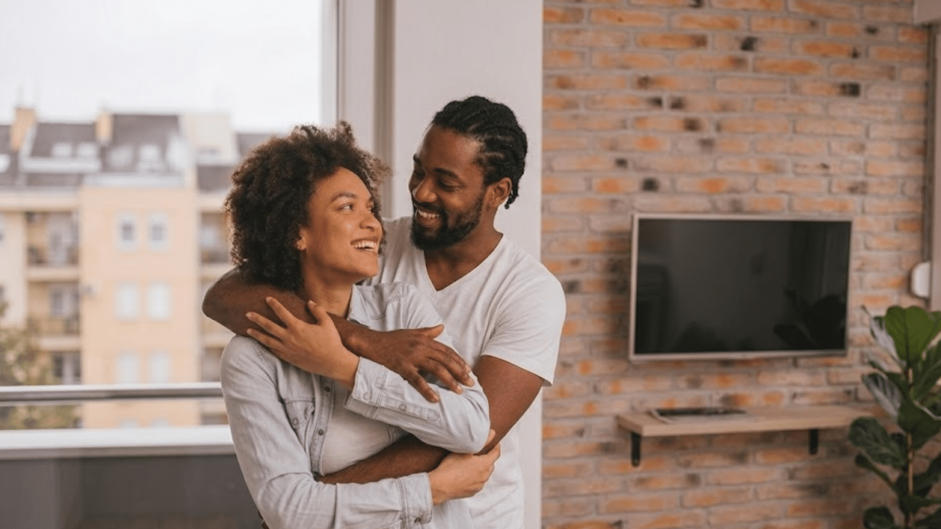 couple embracing and smiling in a bright modern apartment with large windows brick wall and television mounted nearby