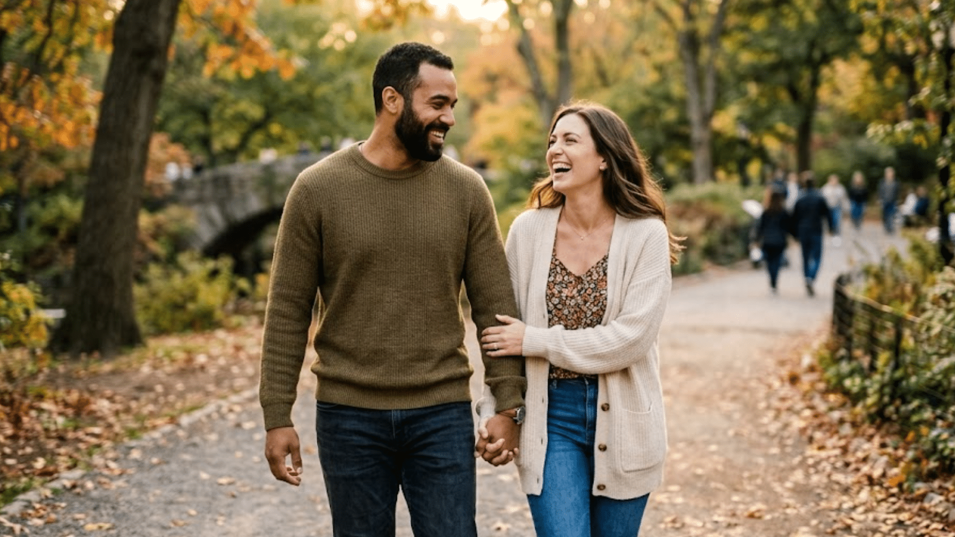 couple holding hands and laughing while walking along a tree lined park path with autumn leaves and people in background