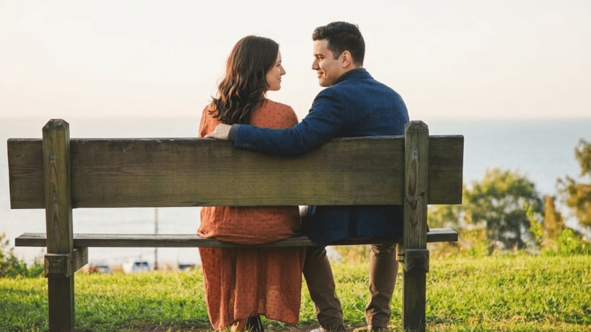 couple sitting on a wooden bench overlooking the water while smiling at each other on a sunny grassy hillside