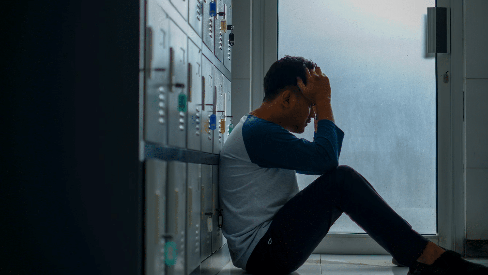 distressed person sits on the floor beside lockers holding his head near a frosted door in dim light