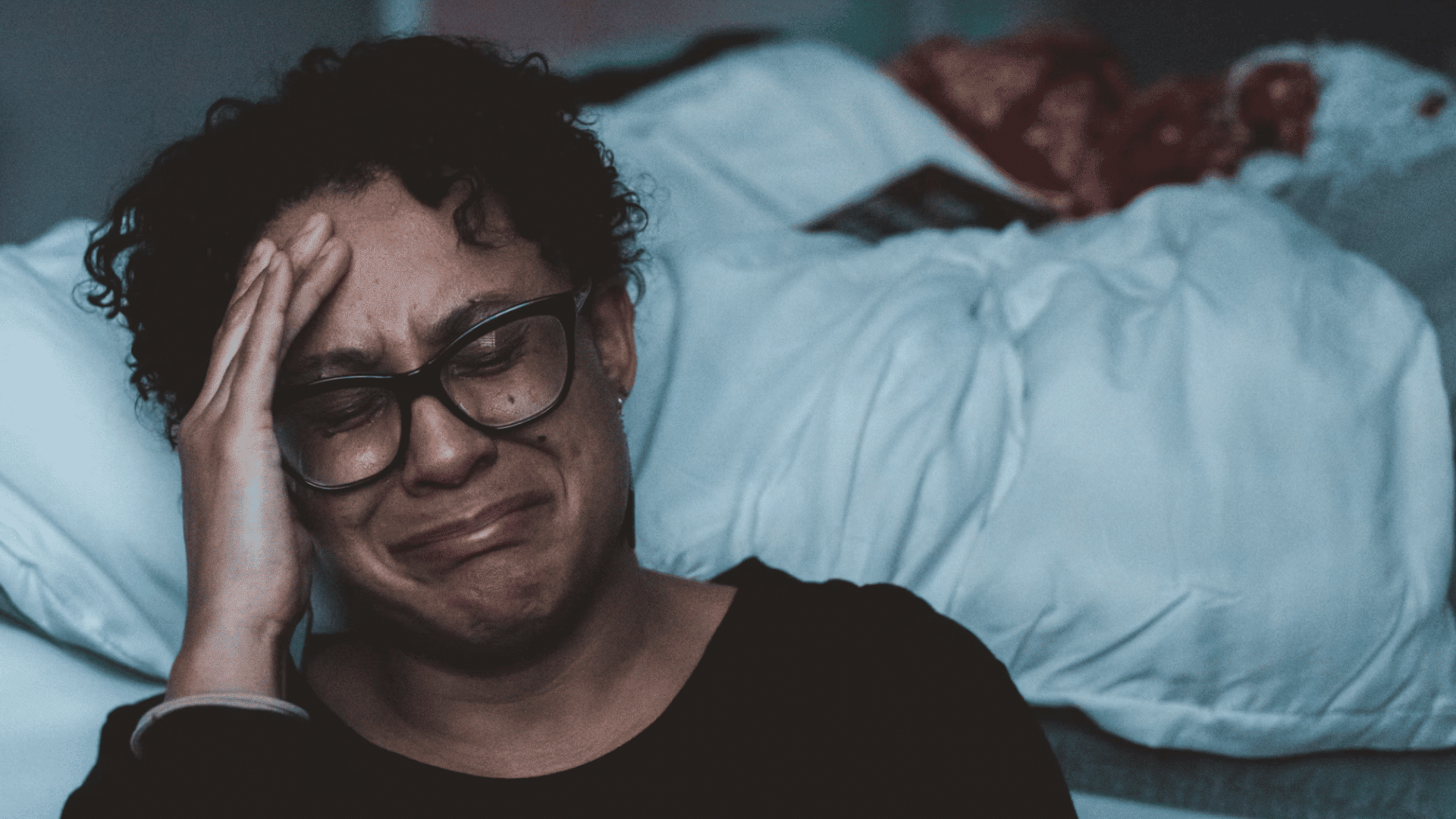 distressed person with glasses crying while holding their hand to their forehead in a bedroom