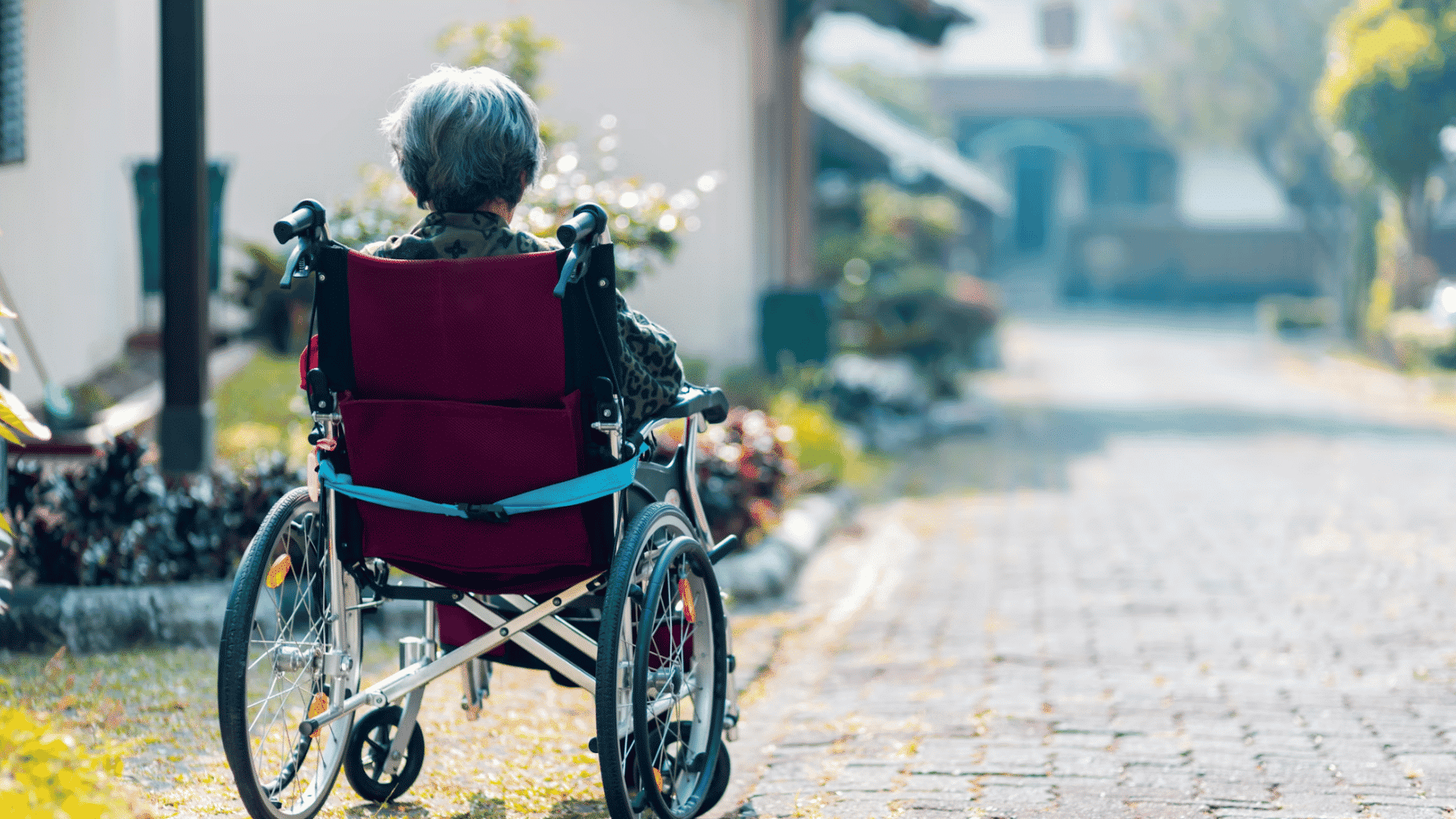 elderly person seated in a wheelchair on a paved path outdoors surrounded by homes and greenery in a quiet neighborhood (1)