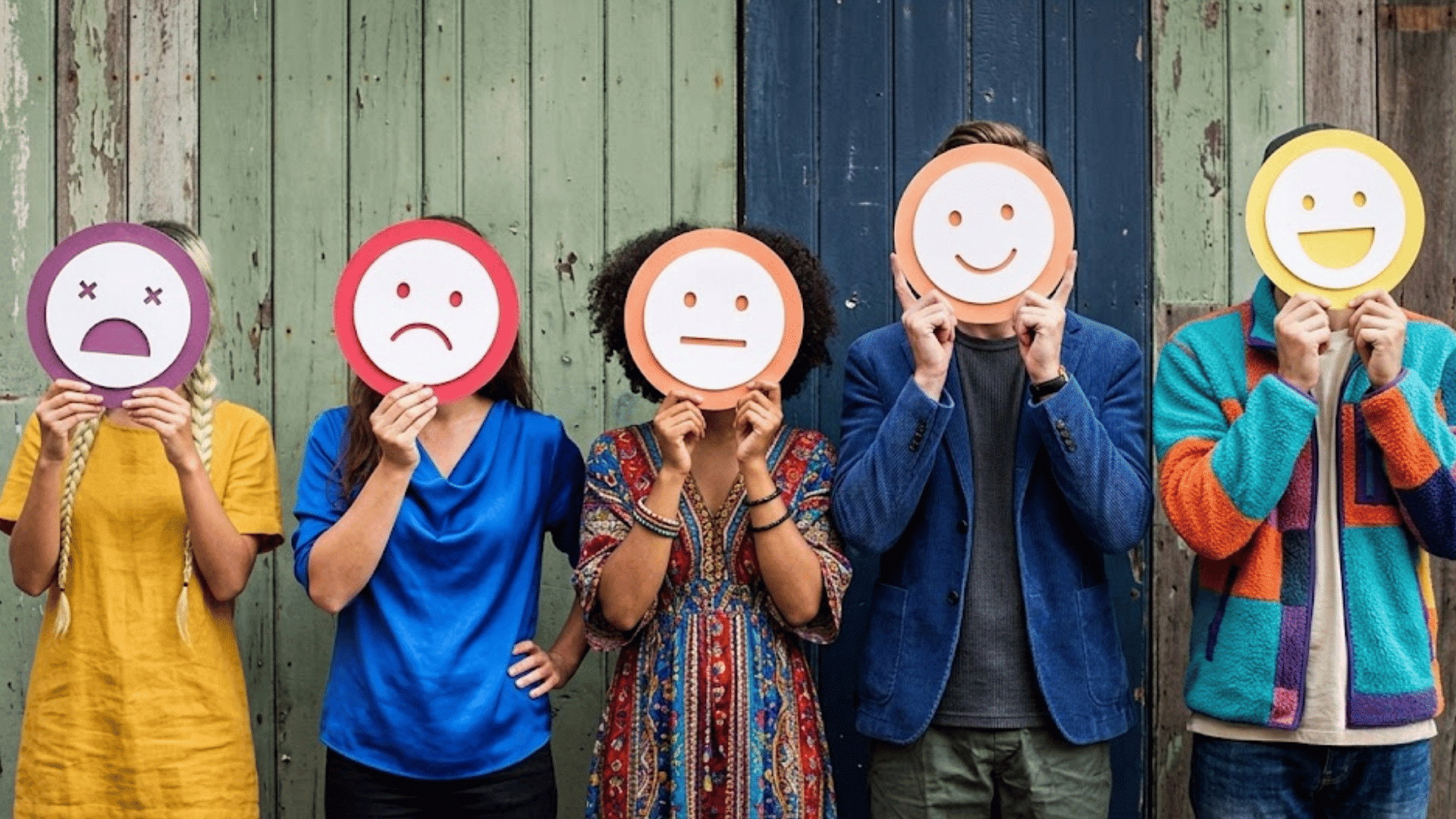 five people standing against a wooden wall holding emoji faces showing sad neutral happy and cheerful moods