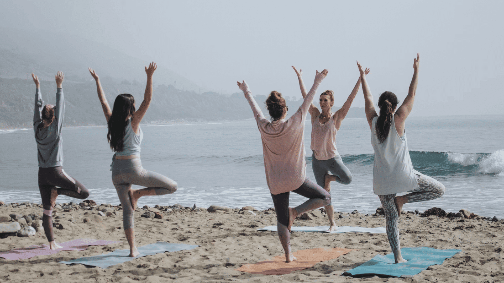 group of people practicing tree pose on yoga mats with raised arms during a beach yoga session beside the ocean