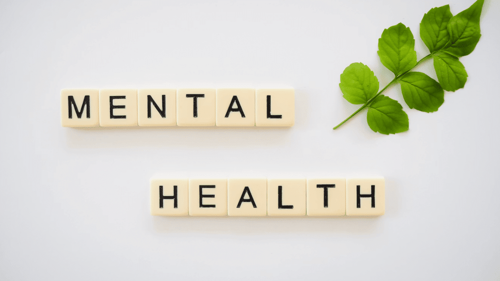 letter tiles spelling mental health beside a green leafy branch on a white background promoting wellbeing awareness