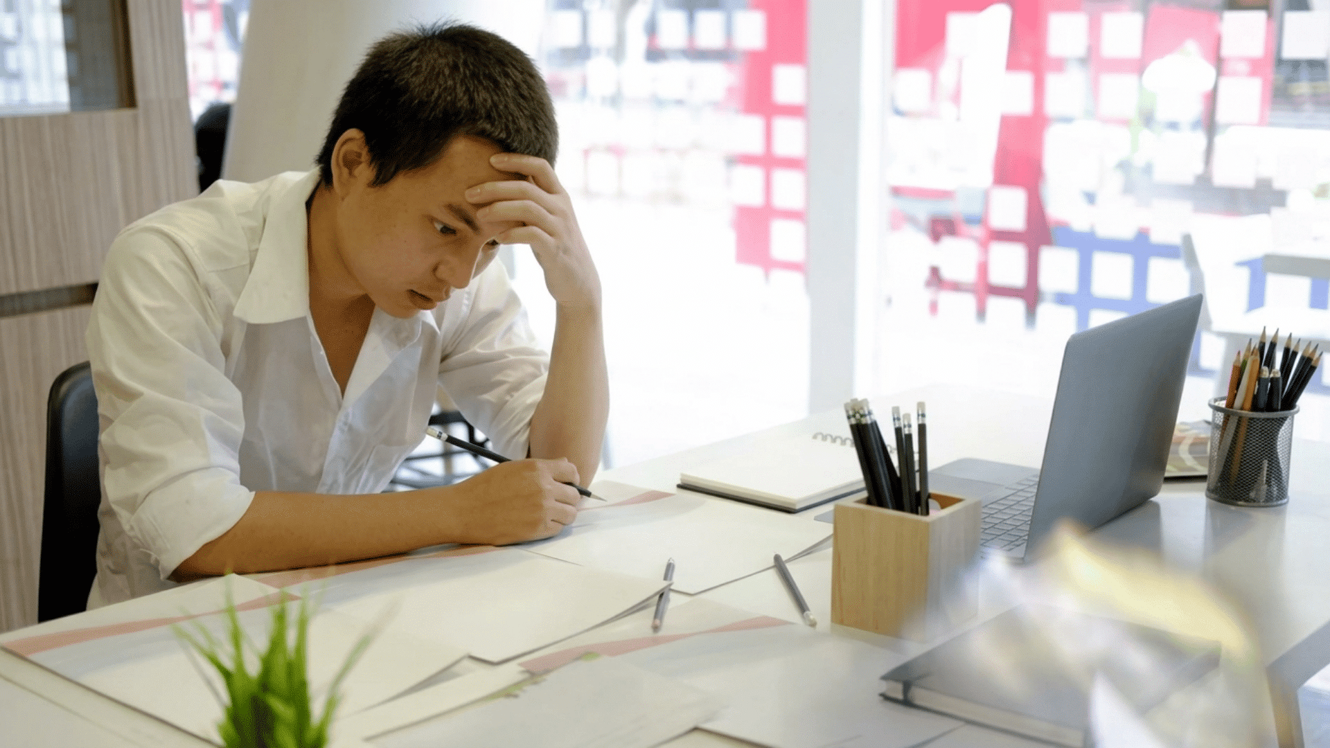 man holding his forehead while writing at a desk beside a laptop and papers