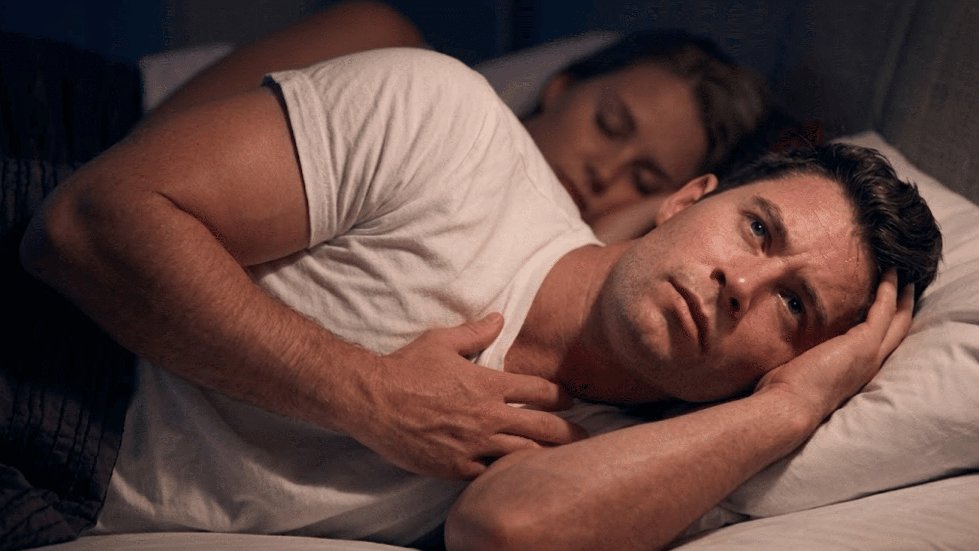 man lying awake in bed looking worried while his partner sleeps in the background depicting relationship anxiety