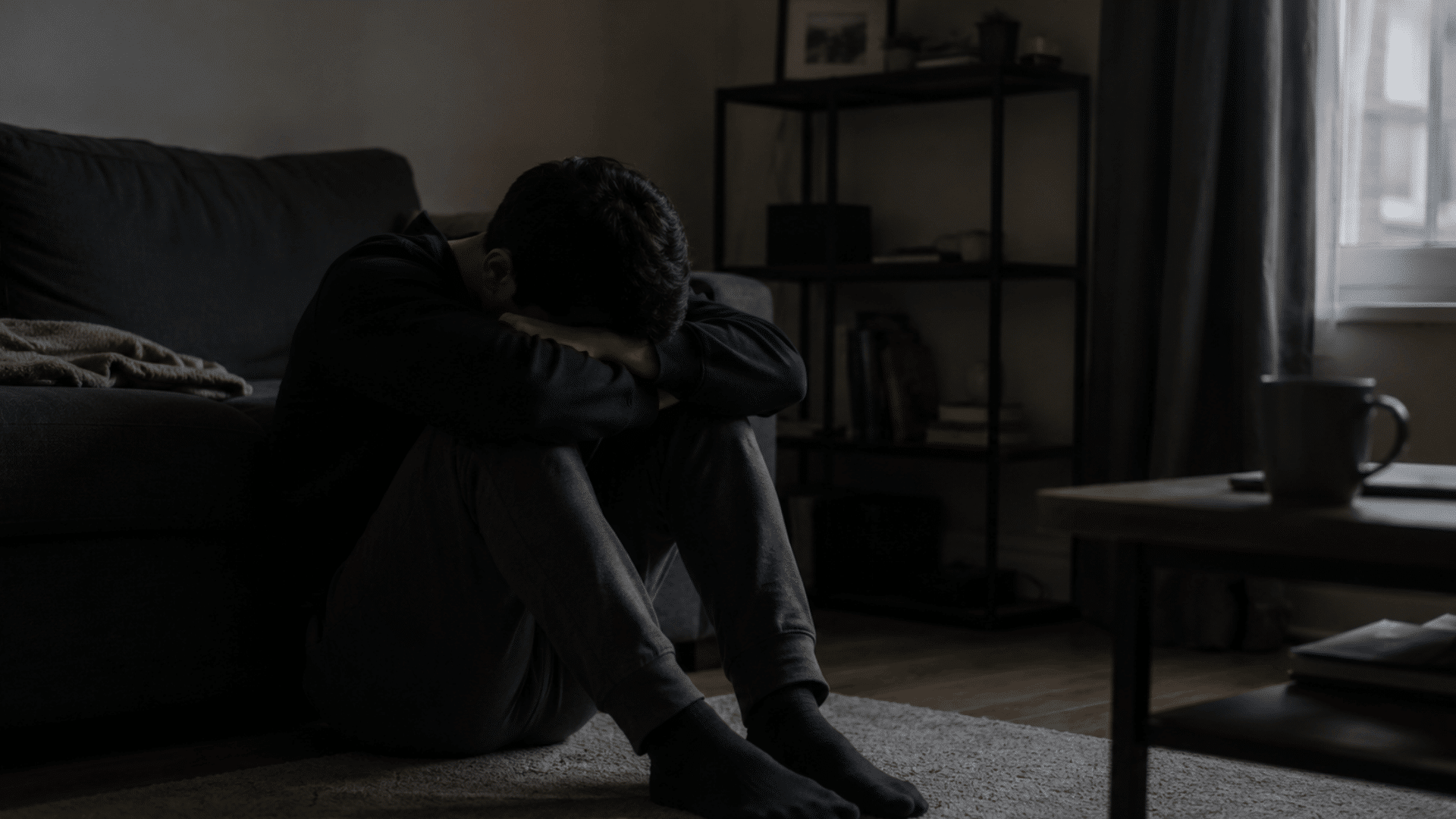 man sits alone on floor beside a sofa with head buried in arms in a dim room showing sadness and distress