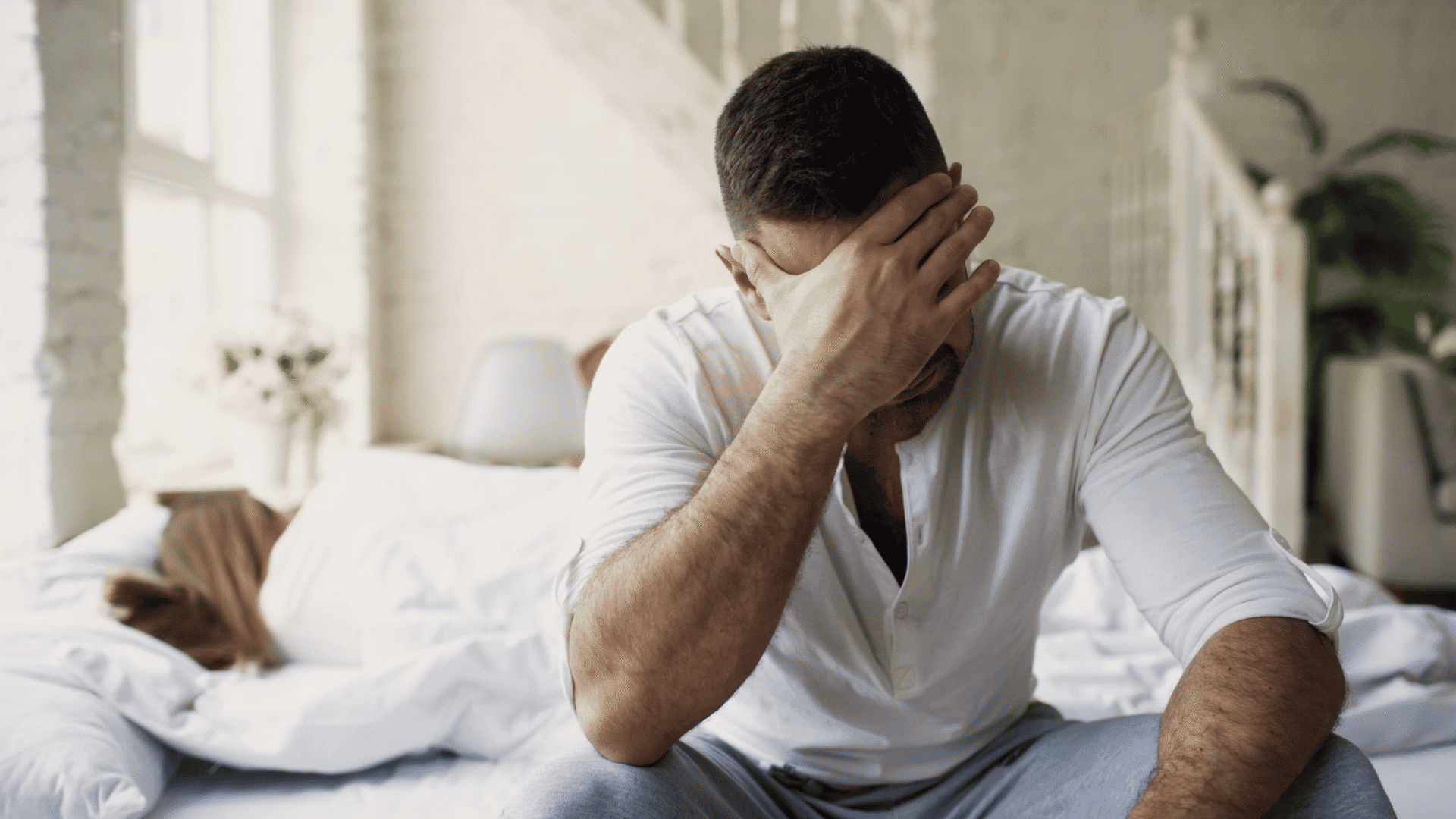 man sits on the edge of a bed covering his face with one hand while appearing distressed in a bright bedroom setting