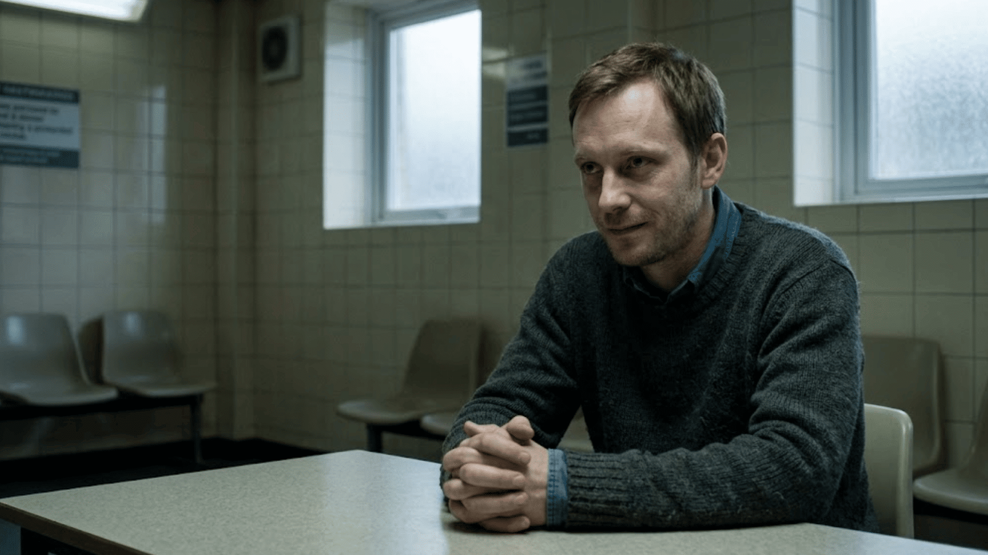 man sitting at a table in a waiting room with hands clasped wearing a dark sweater and looking ahead with a slight smile