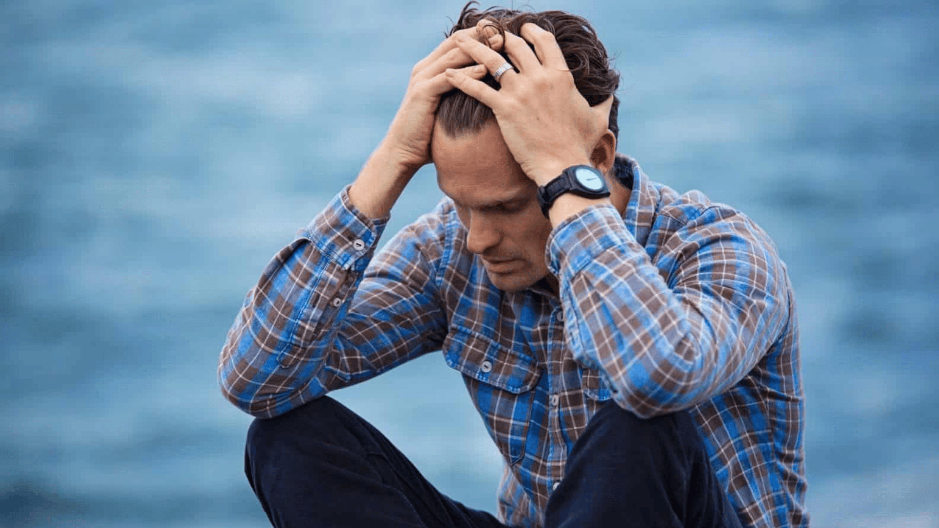 man sitting by the water holding his head showing stress worry and emotional distress outdoors
