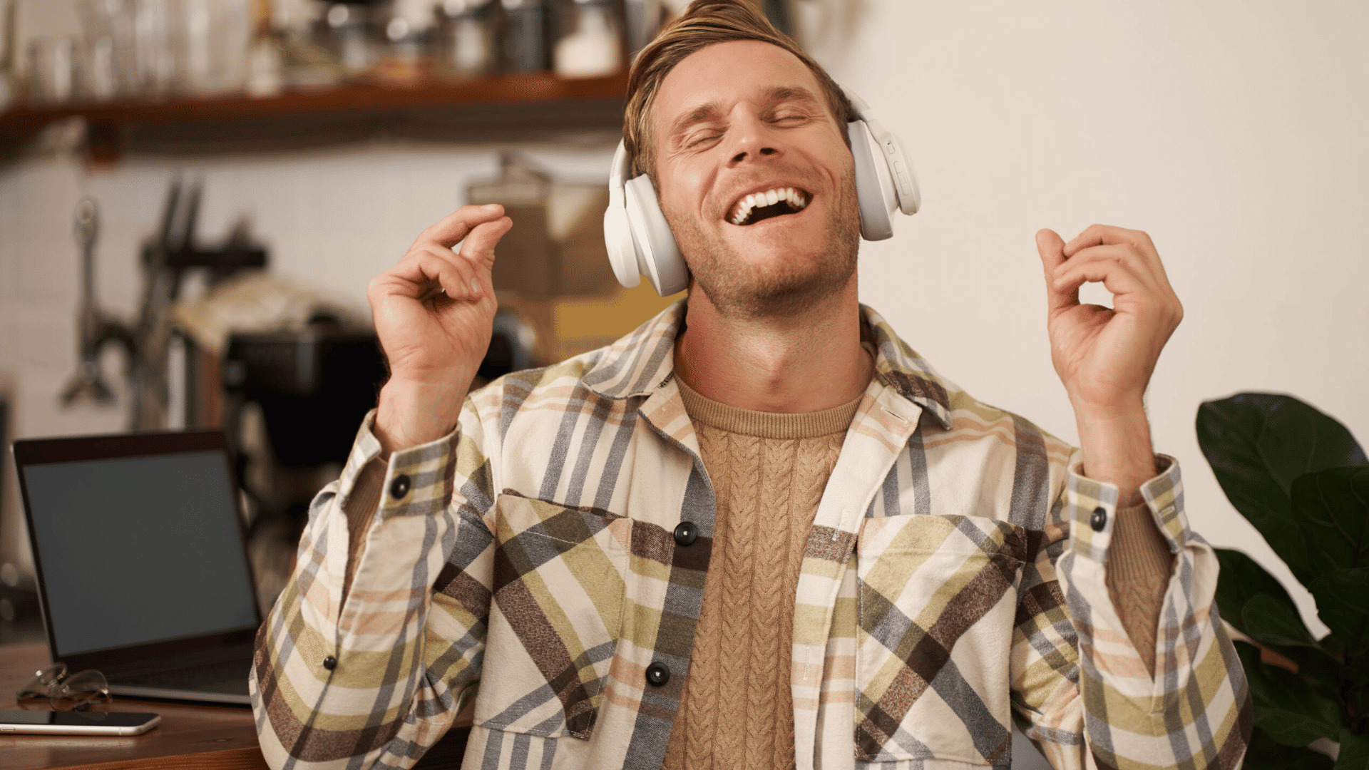 man wearing white headphones singing happily at a desk with laptop and houseplants in the background