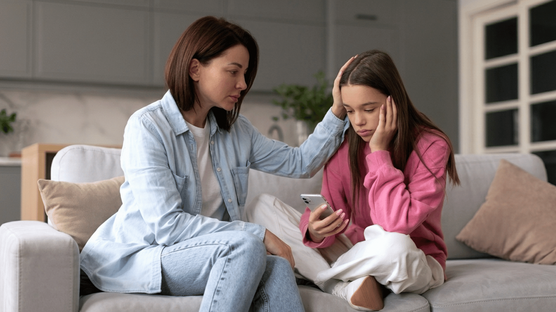 mother comforts sad teen girl holding phone on sofa after feeling rejected by friends at home