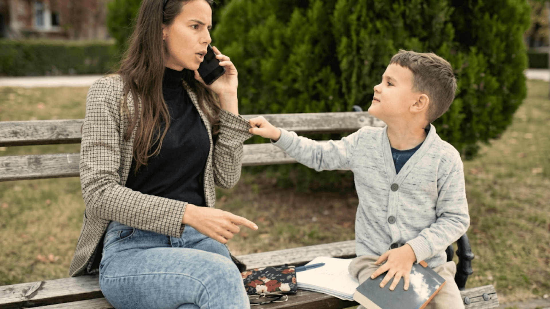 mother talking on phone while child pulls her sleeve seeking attention during time together on a park bench