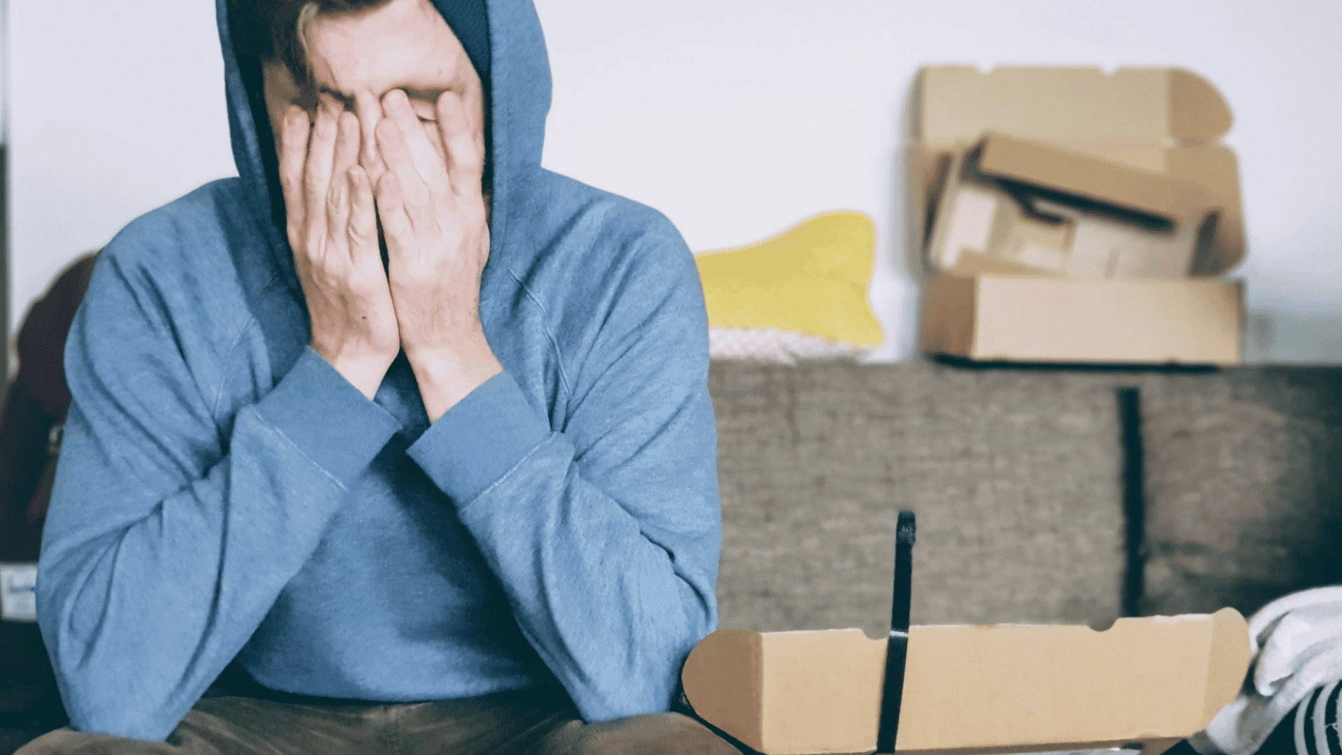 person in a blue hoodie sitting with hands covering face beside opened boxes showing stress during unpacking at home