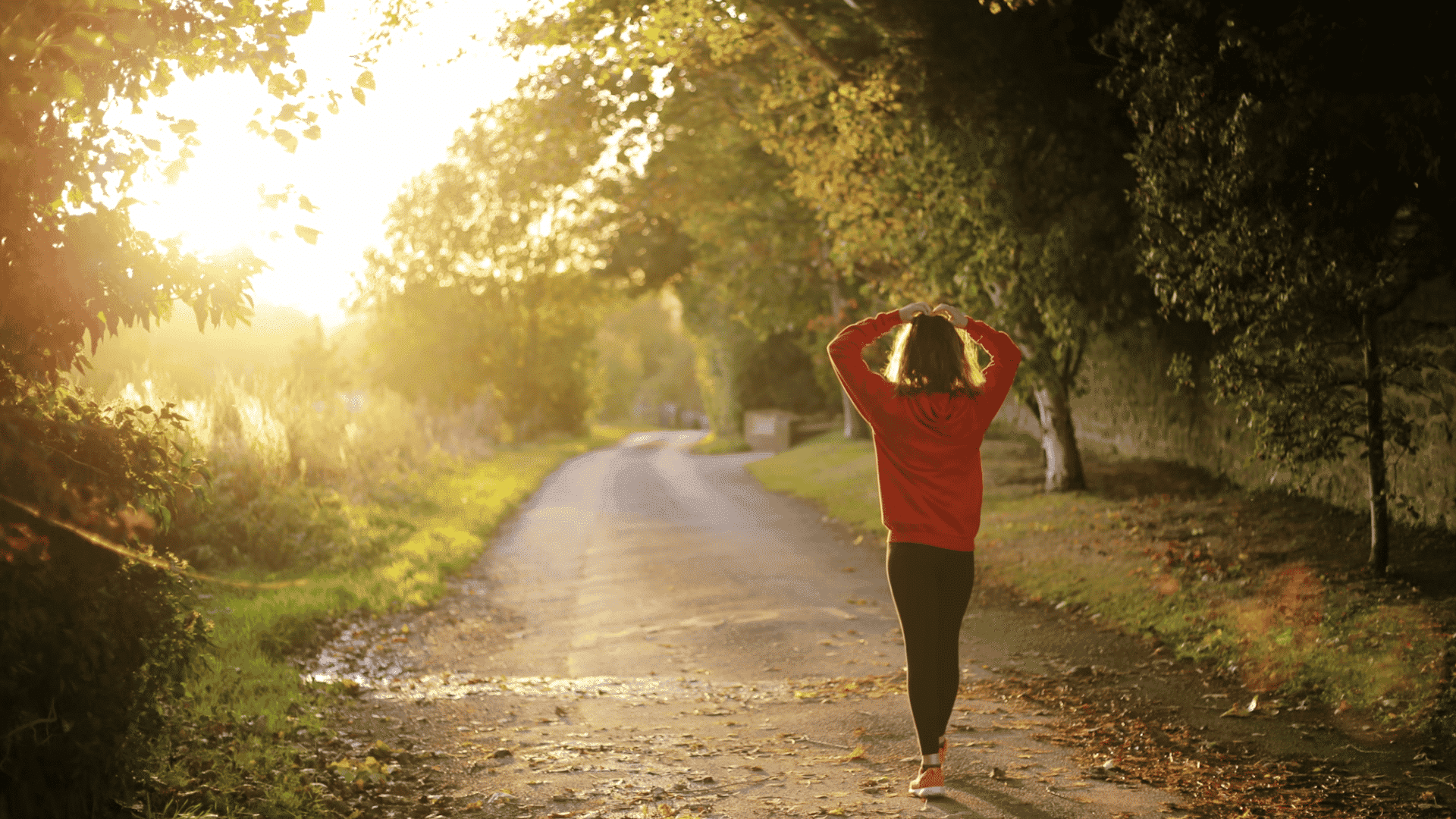 person in a red jacket walking along a sunlit tree lined path during a peaceful outdoor moment
