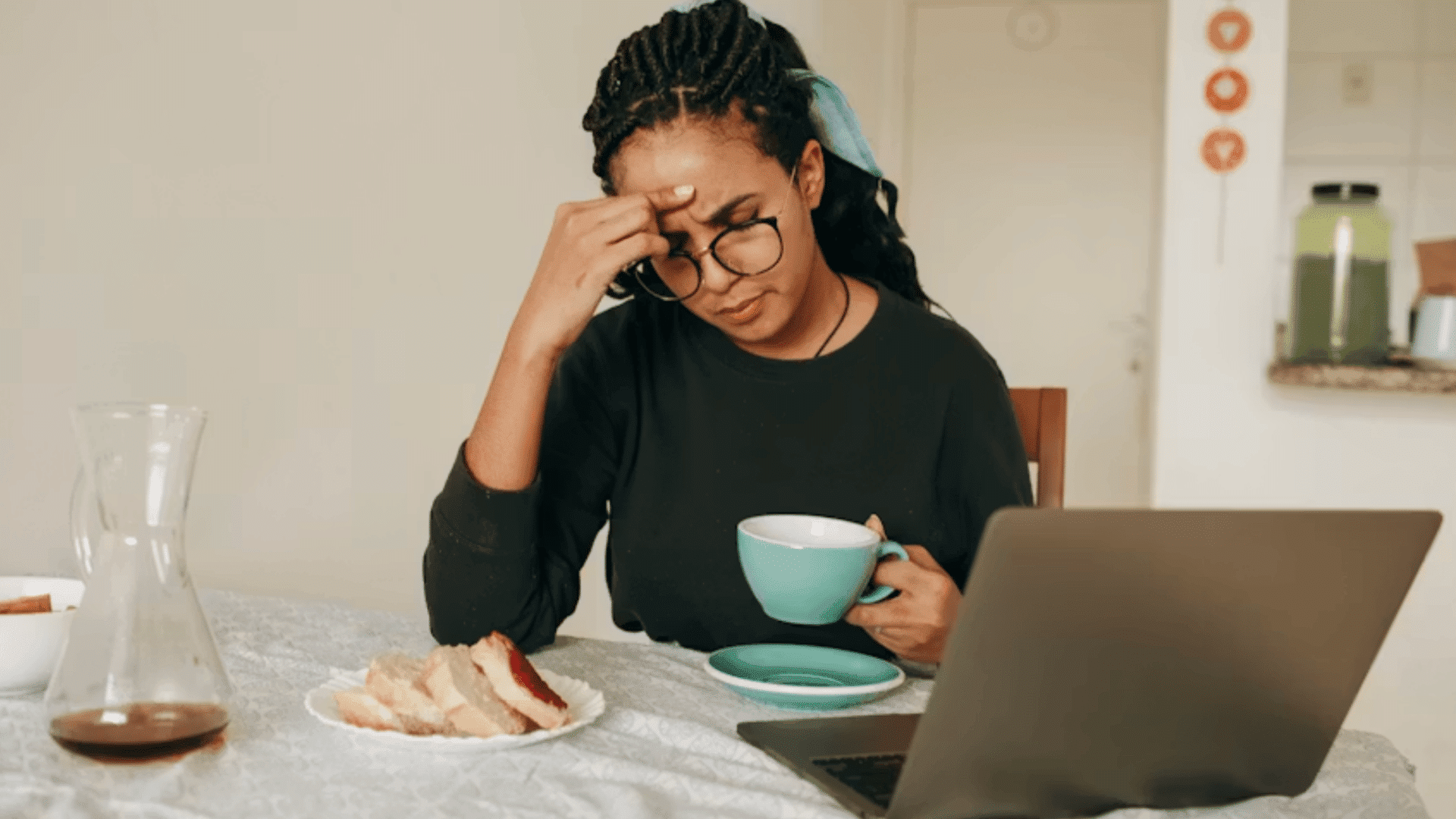 person sitting at a table holding a coffee cup while rubbing their forehead beside a laptop during a stressful work session