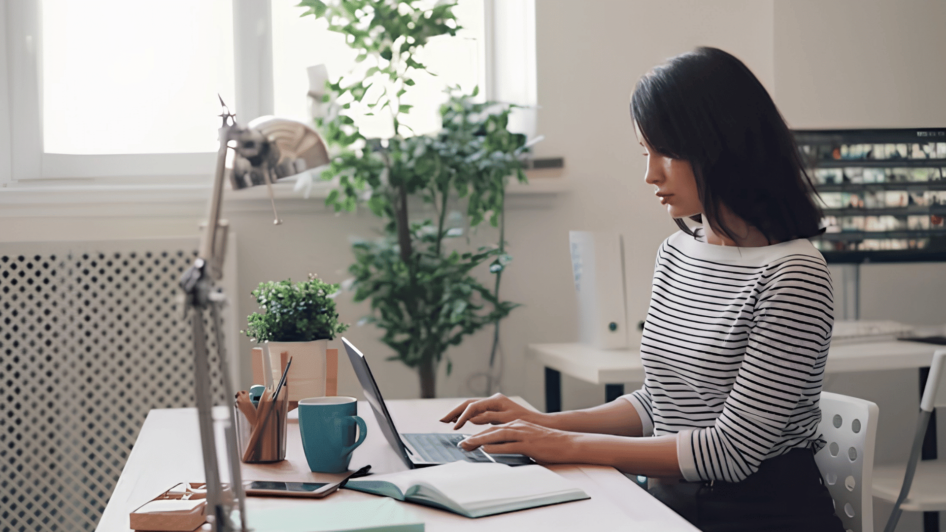 person typing on a laptop at a bright desk with plants and notebooks