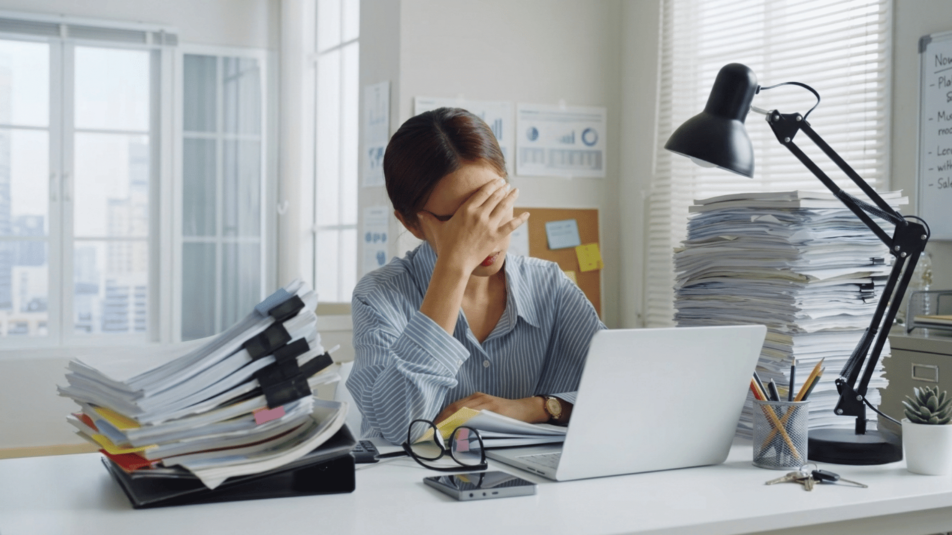 stressed woman sitting at her office desk with a hand on face near laptop and papers