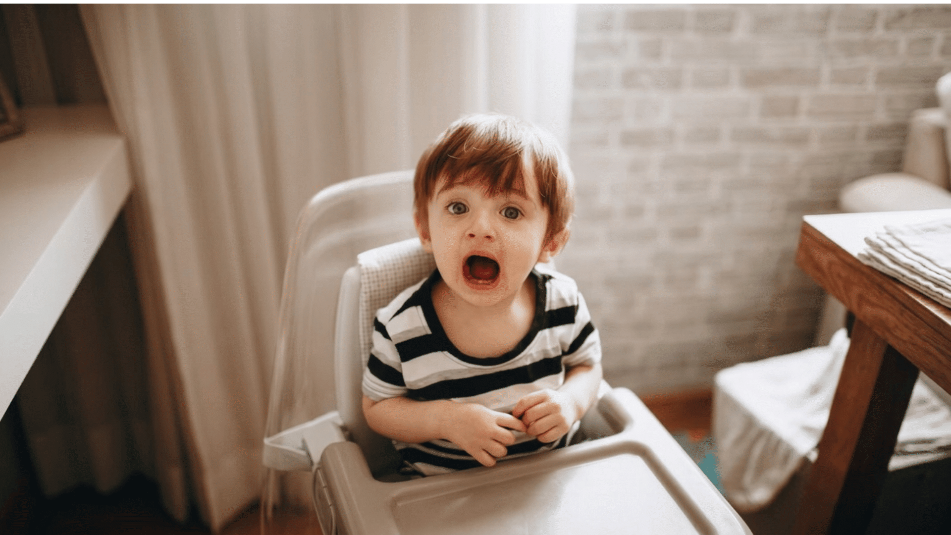 surprised toddler sitting in a high chair with mouth open indoors