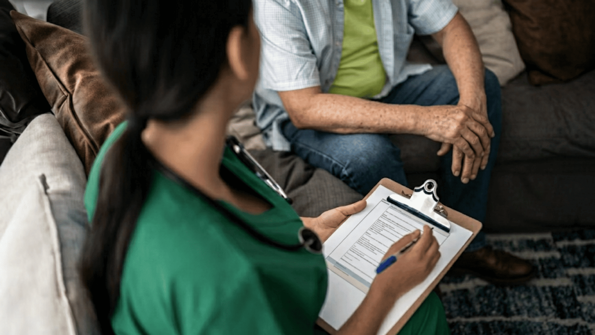 therapist writing on a clipboard during a home session with an older adult seated on a sofa