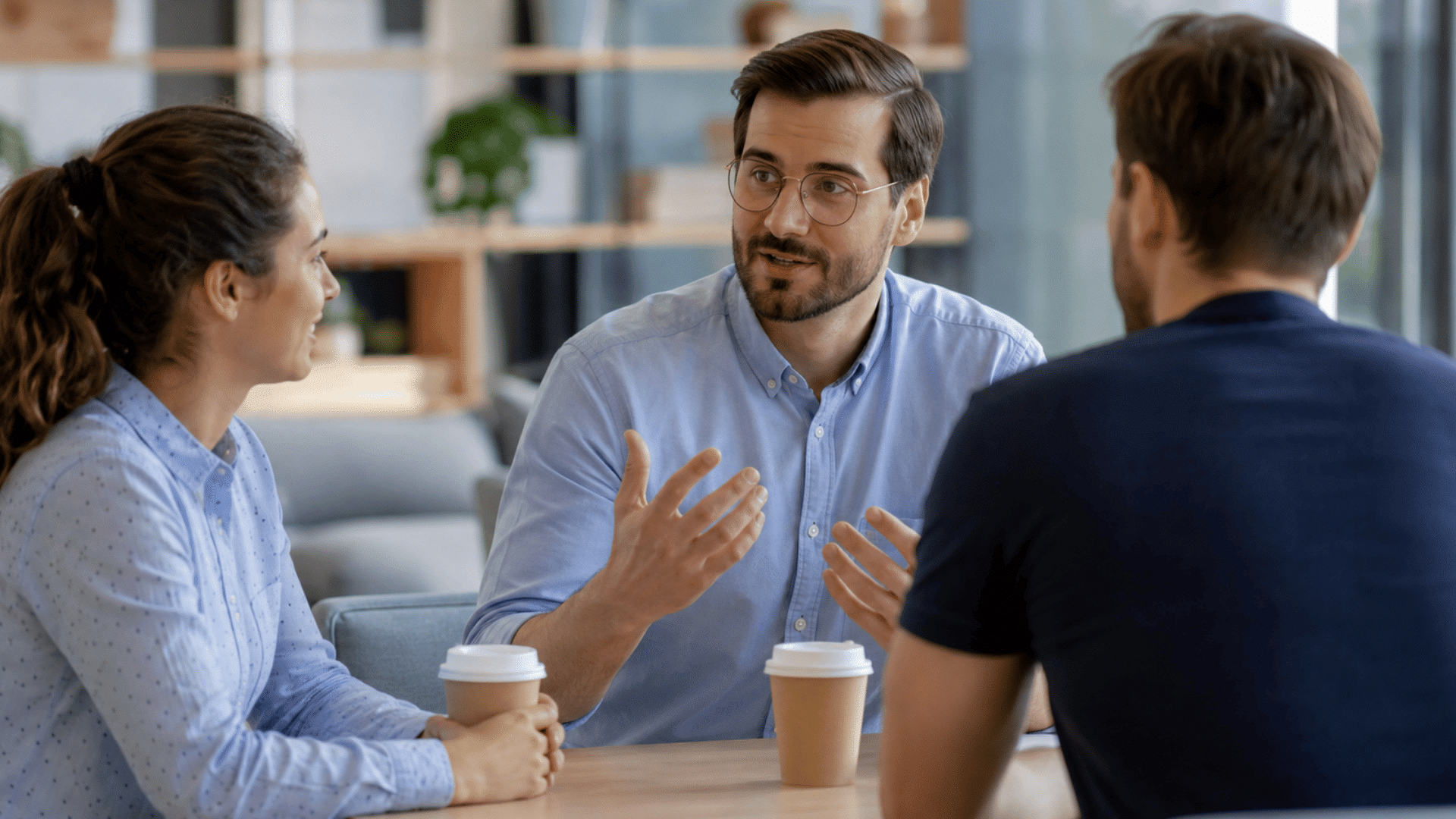 three coworkers sitting around a table sharing coffee while one person gestures during a friendly office discussion