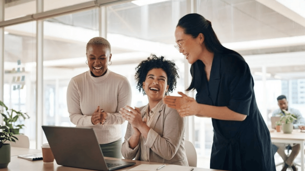 three coworkers smile and applaud around a laptop during a team meeting in a bright modern office workspace